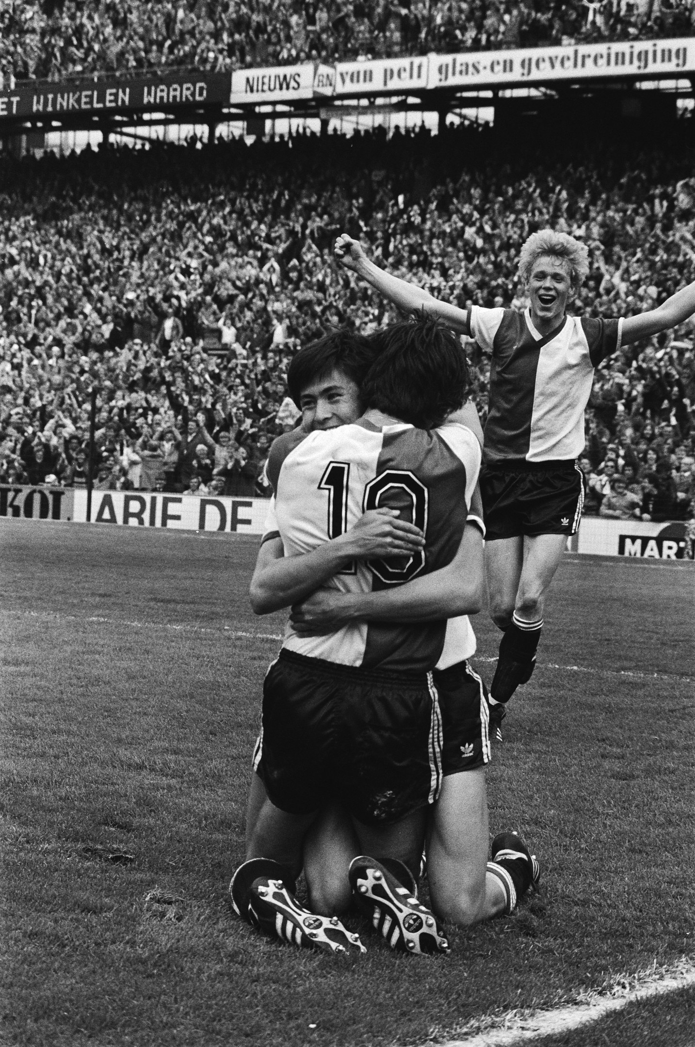 Drei Fußballspieler feiern ein Tor, indem sie sich auf dem Boden umarmen, vor einem jubelnden Publikum und schwenkenden Fahnen im Hintergrund, auf einem Schwarz-Weiß-Bild.