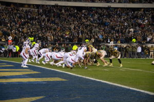 Ein Fußballspiel auf einem Stadionfeld mit Spielern in Sportbekleidung und Helmen, umgeben von Zuschauern und Stadionstrukturen.