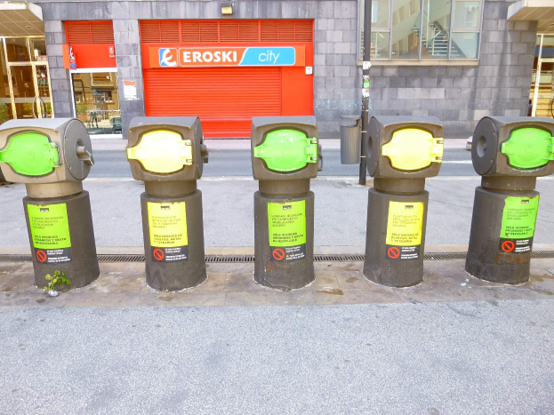 Reihe von Parkautomaten entlang einer Straße mit einem Gebäude mit Glasfenstern und einem Rollladen mit Text im Hintergrund und einem nahen Müllcontainer.
