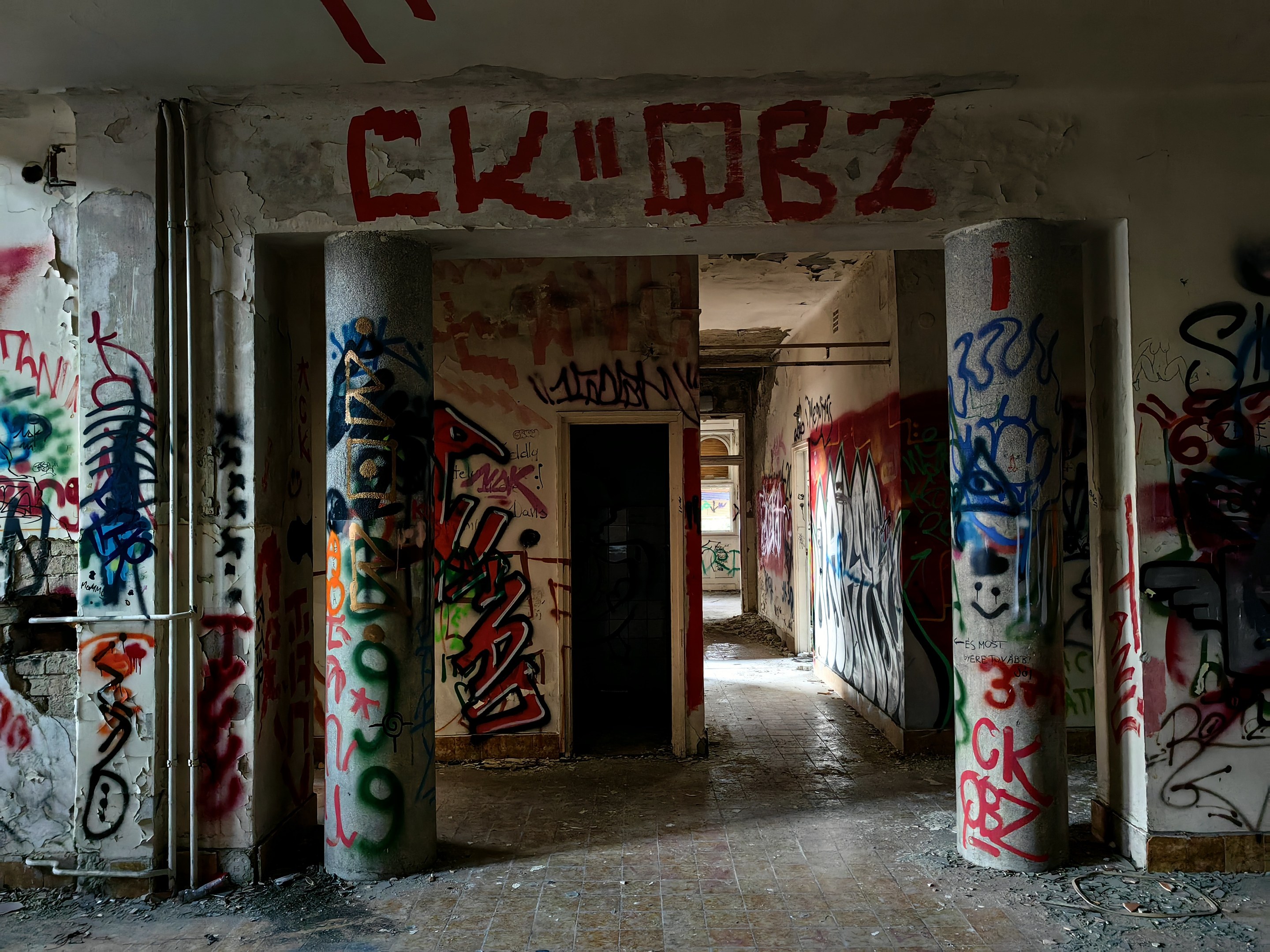 Verlassenes Gebäude mit Graffiti-bedeckten Wänden, Säulen, freiliegenden Rohren und einem sichtbaren Boden, das eine Tür im Hintergrund zeigt.