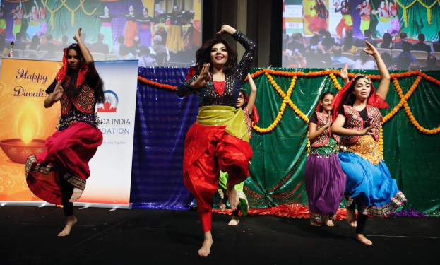 Gruppe junger Mädels, die auf einer Bühne einen traditionellen Tanz aufführen, umgeben von Bannern, Girlanden und einem Publikum im Hintergrund.