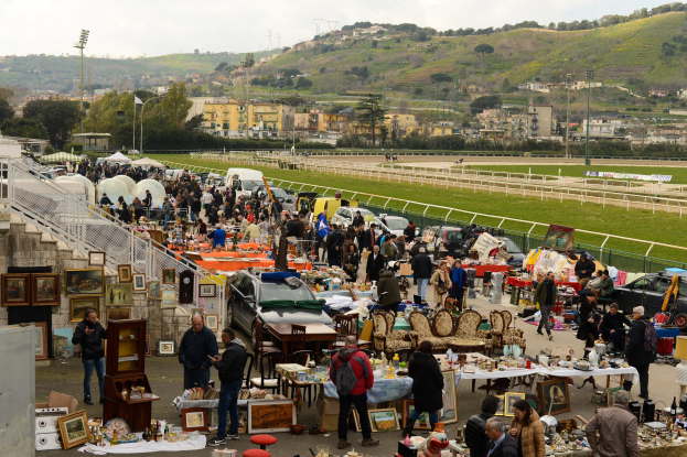 Große Gruppe von Menschen auf einem Flohmarkt mit Tischen voller Gegenstände wie Fotorahmen und Stühle, geparkte Fahrzeuge, Geländer, Stufen, Bäume, Gebäude, Laternenpfähle, Hügel und ein bewölkter Himmel im Hintergrund.