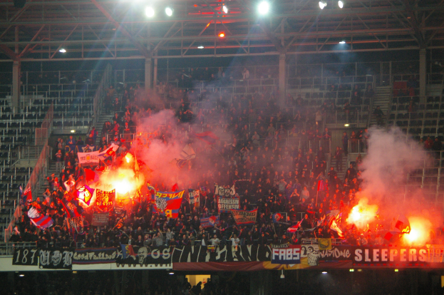 Eine große Menge an Menschen in einem Stadion hält Fahnen und Banner, während Rauch und Bengalos um sie herum aufsteigen, mit Bannern, Metallrahmen, Polen und Deckenleuchten zu sehen.