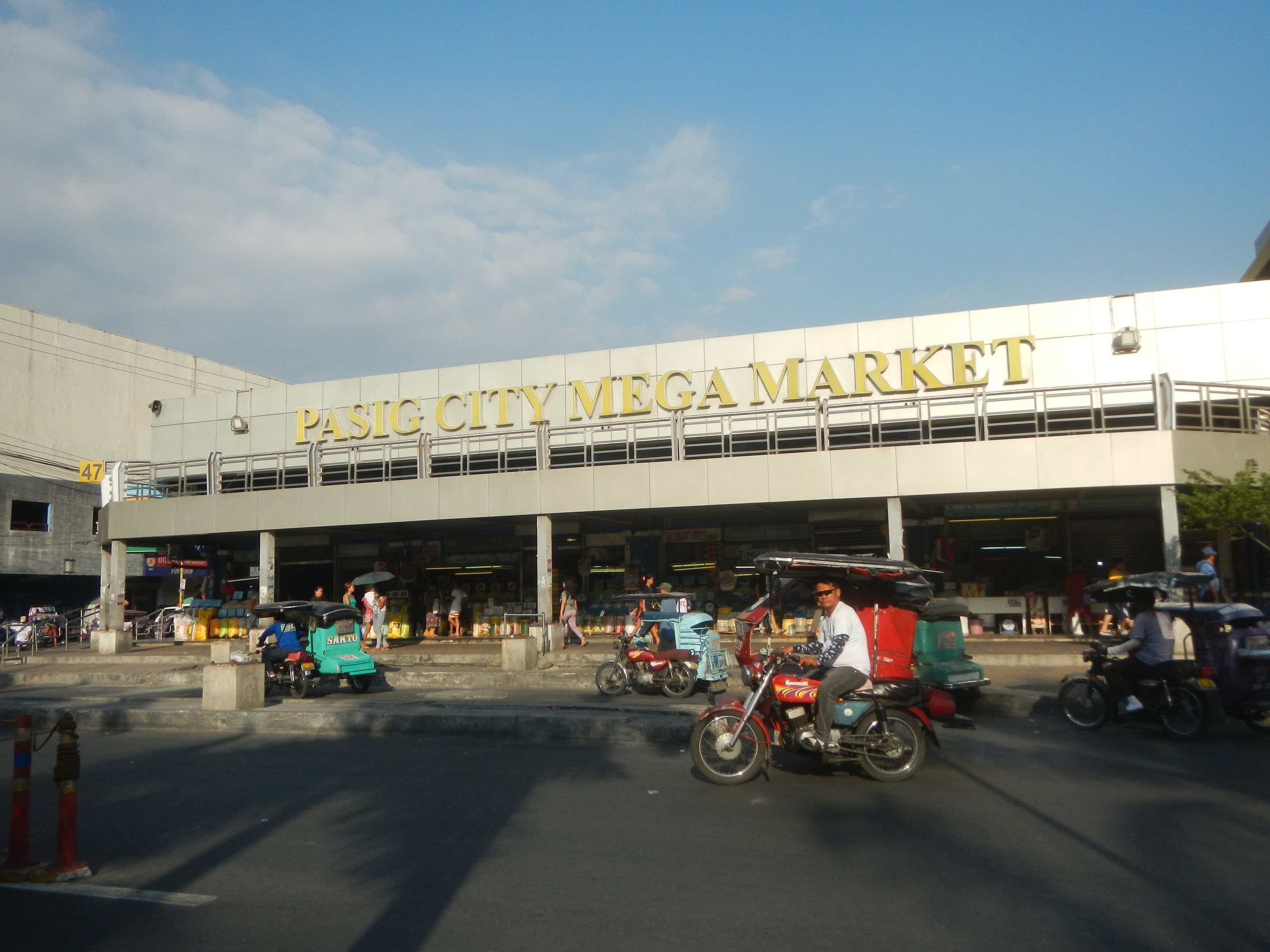 Eine belebte Straßenszene am Pasig City Mega Market mit Fahrzeugen und Fußgängern, ein Gebäude mit Text im Hintergrund und ein bewölkter Himmel.