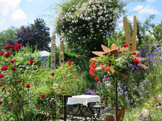 Ein Garten mit bunten Blumen und Pflanzen, der einen Tisch mit einer Decke und Stühlen zeigt, vor Bäumen und einem bewölkten Himmel auf dem Chelsea Flower Show.