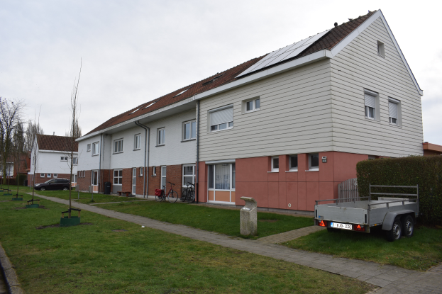 Ein Lastwagen steht vor einer Reihe von Häusern mit Solarpaneelen auf dem Dach, Fahrräder in der Nähe und ein Weg, der zu den Häusern führt, mit Pflanzen, Bäumen und einer bewölkten Landschaft im Hintergrund.