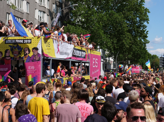Eine große Menge marschiert während des Berlin Pride 2019 die Straße entlang, viele tragen Kappen und Goggles, einige halten Fahnen, mit Bannern, Laternenpfählen, Bäumen und Gebäuden im Hintergrund unter einem bewölkten Himmel.