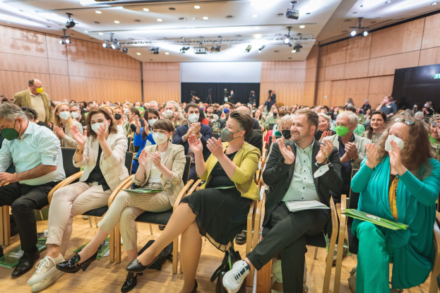 Eine Gruppe von Menschen in Stühlen klatschend, einige tragen Masken, mit Taschen auf dem Boden, vor einer Menge bei einer Coronavirus-Konferenz mit einem Bildschirm und Deckenlampen im Hintergrund.
