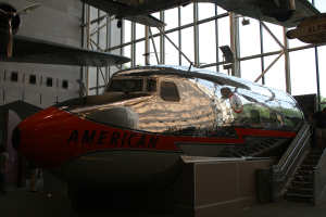 Ein Flugzeug von American Airlines in einem Museum ausgestellt, mit Menschen auf dem Boden, einer Treppe auf der rechten Seite, die zum Flugzeug führt, und einem Gebäude mit Fenstern, Bäumen und einem klaren blauen Himmel im Hintergrund.