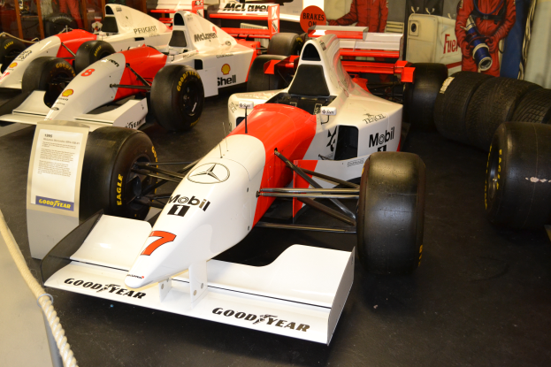Eine Gruppe von Rennwagen im Museum ausgestellt, umgeben von Menschen, ein Schild, eine Absperrung und eine Wand im Hintergrund.