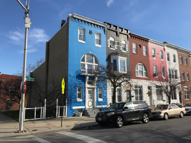 Eine Reihe bunter Häuser an einer Straßenecke in Baltimore, Maryland, mit parkenden Autos, Bäumen, Strommasten und einem Zaun unter einem bewölkten Himmel.