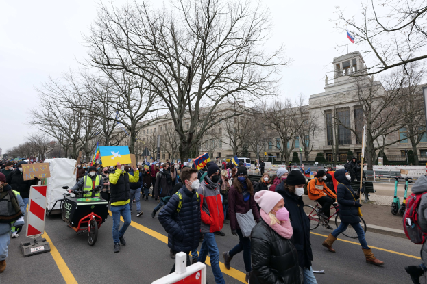 Eine große Gruppe von Menschen bei einer Demonstration auf einer Straße in Washington, D.C., mit Schildern und Transparenten, einige fahren Fahrräder, unter einem klaren blauen Himmel.