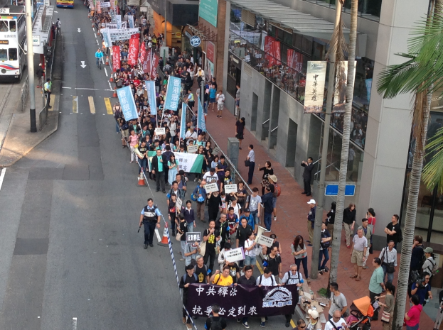 Eine große Gruppe von Menschen marschiert auf einer Straße in Hong Kong, mit Transparenten und Schildern, mit Bäumen, Glasgebäuden, Fahrzeugen und Straßenschildern im Hintergrund.