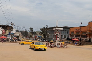 Eine belebte Stadtstraße mit einem gelben Taxi, das fährt, Fußgänger mit Schirmen auf den Gehwegen, Gebäude mit Fenstern und Strommasten mit Drähten unter einem bewölkten Himmel.