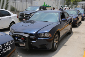 Polizeifahrzeuge auf einem Parkplatz geparkt mit Menschen auf der Straße, umgeben von einem Zaun, Bäumen, Gebäuden und einem klaren blauen Himmel.