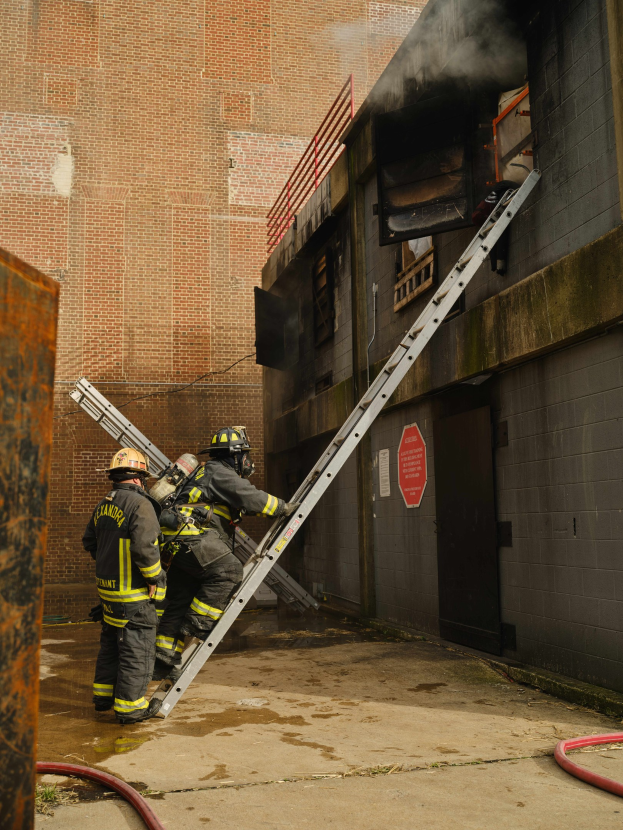 Feuerwehrleute in Helmen, die eine Leiter zu einem raucherfüllten Gebäude hinaufsteigen, mit einem metallischen Objekt links und Rohren an der Basis.