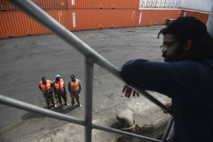 Ein Mann in legerer Kleidung steht auf einem Metallgeländer in der Nähe einer Gruppe von Menschen in orangefarbenen Jacken und Mützen, mit Containern und einer Wand im Hintergrund am Hafen von Los Angeles.