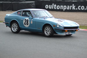 Blauer Wagen auf Rennstrecke mit Helmträger am Steuer, umgeben von Reifen, einem Banner mit Text und grasbewachsenem Boden.