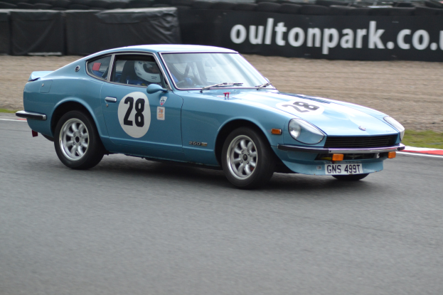 Blauer Wagen auf Rennstrecke mit Helmträger am Steuer, umgeben von Reifen, einem Banner mit Text und grasbewachsenem Boden.