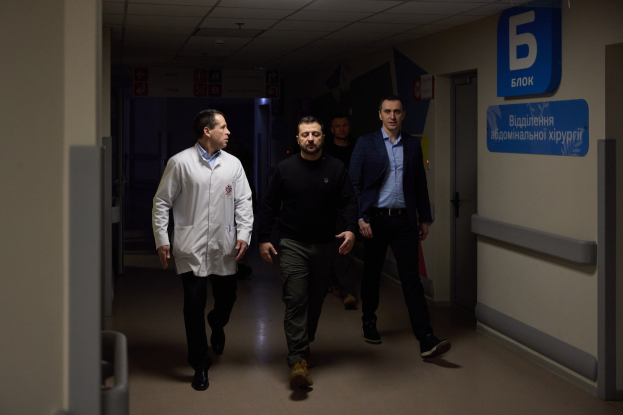 Eine Gruppe von Männern geht einen Krankenhausflur entlang mit Deckenbeleuchtung, einer Tür an der rechten Wand und Texttafeln an der linken Wand, die besorgt aussehen.