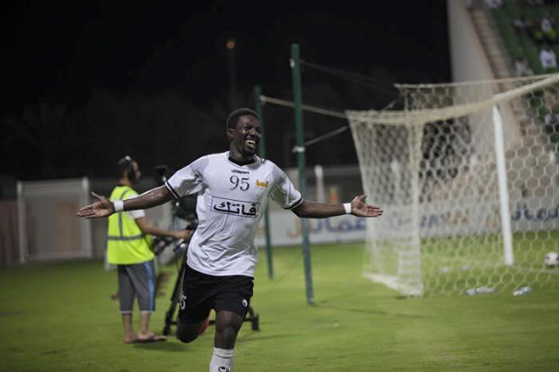 Mann in weißem Hemd und schwarzen Shorts rennt auf einem Fußballfeld mit ausgestreckten Armen, feiernd, umgeben von Menschen, mit einem Tor auf der rechten Seite und Zuschauern im Hintergrund unter einem dunklen Himmel.