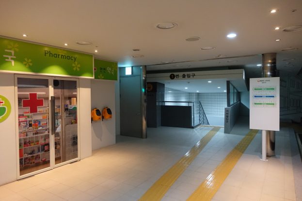 Apothekeneingang mit einem "Apotheke"-Schild auf der linken Seite, einer Tafel mit Text auf der rechten Seite, beleuchteten Lampen auf dem Dach und undeutlichen Hintergrundobjekten.