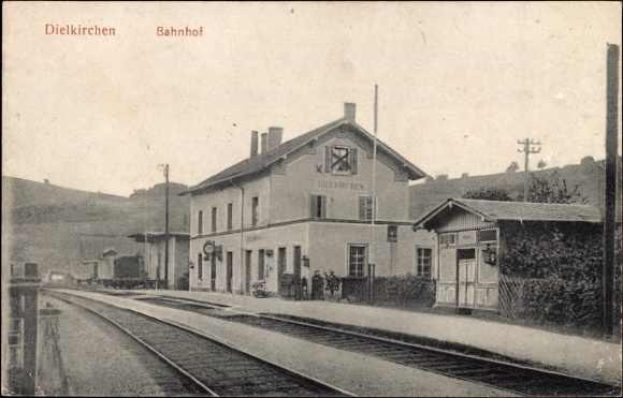 Schwarzes und weißes Foto einer Bahnhofstation, die als "Dielkirchen, Bahnhof, Deutschland" beschriftet ist, mit einem Zug auf den Gleisen, umgeben von Häusern, Strommasten, Bäumen, Menschen und Bergen sowie einem klaren Himmel.