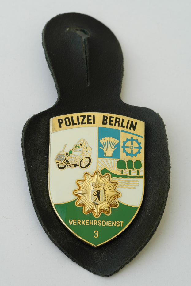 Ein schildförmiges Polizeisiegel mit 'Polizei Berlin' in schwarzer Schrift auf einem blauen Hintergrund mit einem weißen Stern und Rahmen, das auf einer Oberfläche ruht.