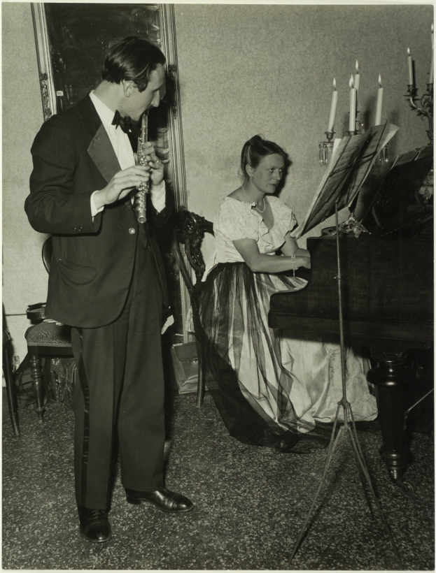 Ein Mann im Smoking spielt Klarinette, während eine Frau an einem Piano sitzt und Noten auf dem Ständer hat, gerahmtes Kunstwerk ist an der Wand im Hintergrund zu sehen.