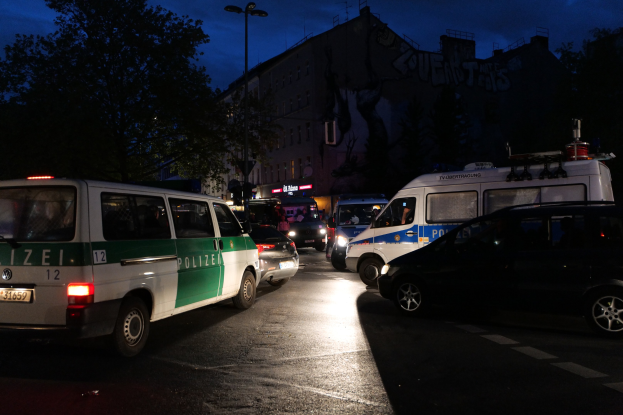 Eine Gruppe von Polizeiwagen, darunter ein Van, ein Auto und ein Rad, parkten am Straßenrand bei Nacht, mit Gebäuden, Bäumen, Laternenpfählen und einem Sternenhimmel im Hintergrund.
