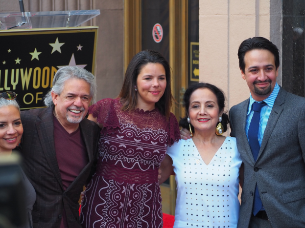 Gruppe von Menschen auf einem roten Teppich bei der Zeremonie des Hollywood Walk of Fame posierend, mit einer Kamera auf der linken Seite, einer Tafel mit Text, einem Glaspodest und einer Wand im Hintergrund.