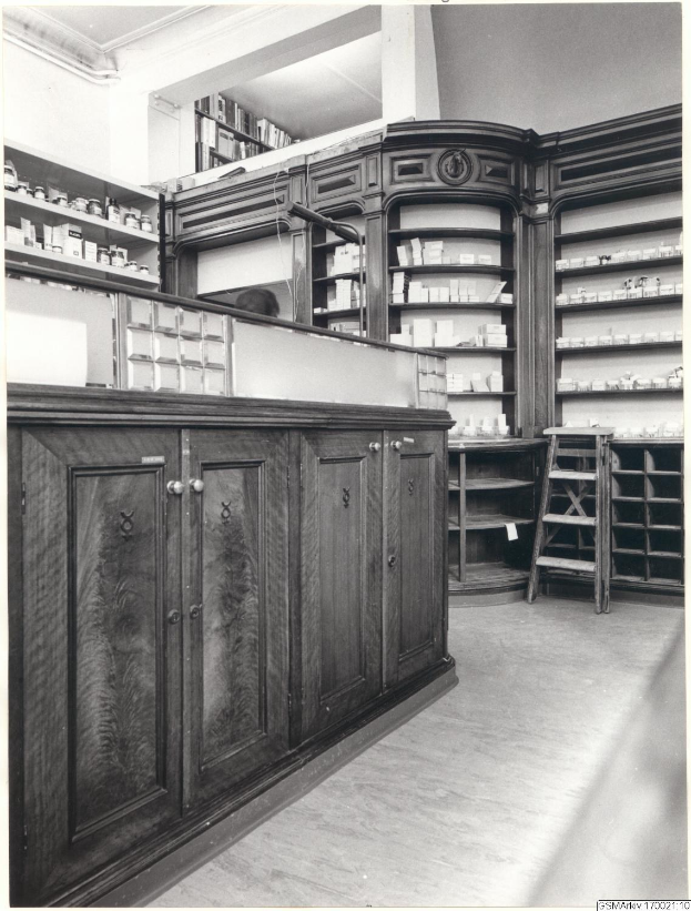 Ein Schwarz-Wei├č-Foto eines Apothekeninneren mit einer Theke, Sch├╝ben, einer Leiter und Regalen, die mit verschiedenen Gegenst├Ąnden gef├╝llt sind, mit Text ├╝ber dem Bild.