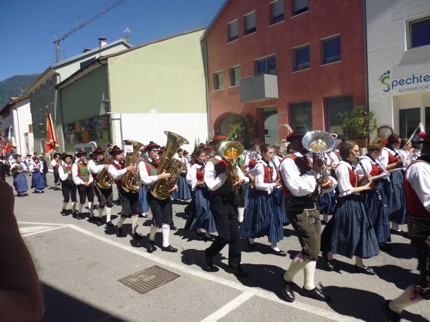 Eine Gruppe von Menschen in traditioneller bayrischer Tracht, die auf einer Straße mit Gebäuden Musikinstrumente spielen und einige Fahnen halten, mit einem Hügel und einem blauen Himmel im Hintergrund.