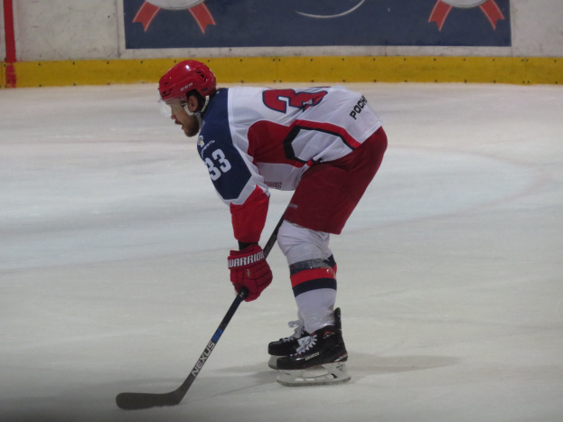 Ein Hockey-Spieler in voller Ausrüstung fährt auf dem Eis, hält einen Stock und hat ein Banner mit Text und Logos im Hintergrund.