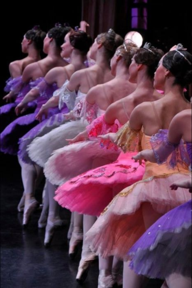 Balletttänzerinnen in rosafarbenen Tutus sitzen auf einer Bühne, beleuchtet von einem Licht im Hintergrund, bewegen sich synchronisiert.