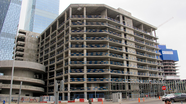 Großes Bauprojekt in einer Stadt mit Wolkenkratzern, Fahrzeugen, Fußgängern, Verkehrsinfrastruktur und Kränen vor einem klaren blauen Himmel.