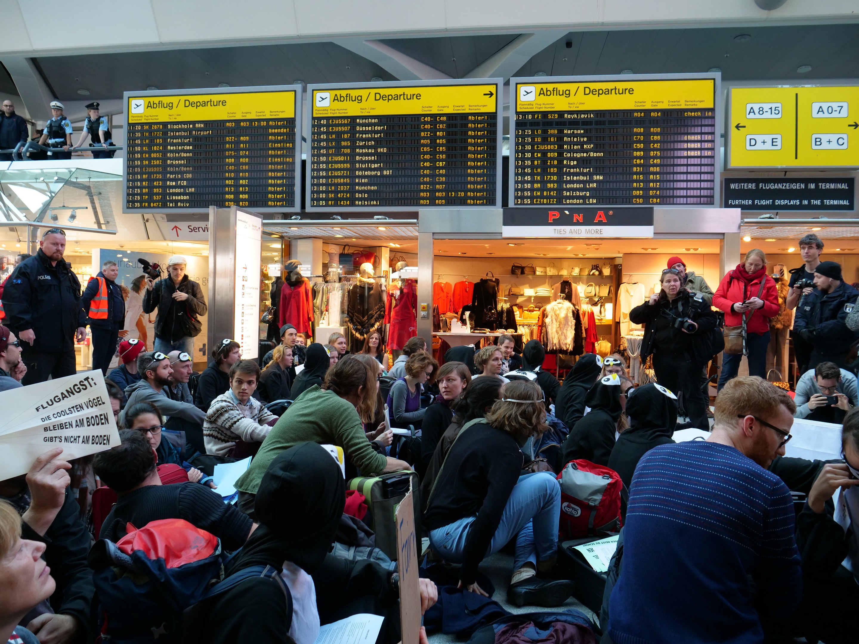 Eine große Gruppe von Menschen sitzt und steht in einem Flughafen während einer Demonstration, mit Informationsschildern, Puppen in Kleidern und Deckenbeleuchtung im Hintergrund.