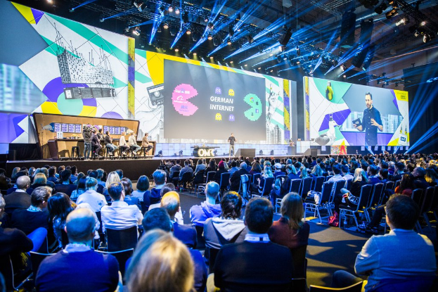 Eine große Menschenmenge sitzt auf Stühlen vor einer Bühne bei der Deutschen Internet-Konferenz 2019, mit einem Tisch voller Gegenstände links und Bildschirmen und Lichtern im Hintergrund.