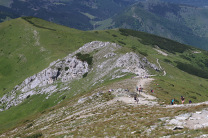 Gruppe von Menschen beim Wandern auf einem Berghang mit grünem Gras und felsigem Gelände, Himmel im Hintergrund sichtbar