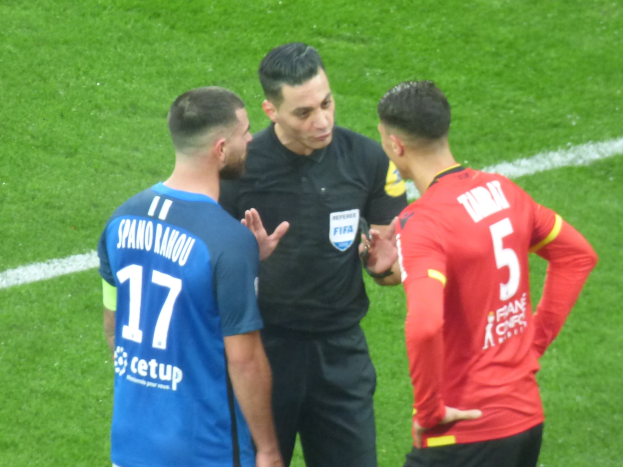 Schiedsrichter spricht mit zwei Männern auf einem Fußballfeld; einer trägt schwarz, der andere ein blau-rotes T-Shirt mit Text, beide tragen Brillen.