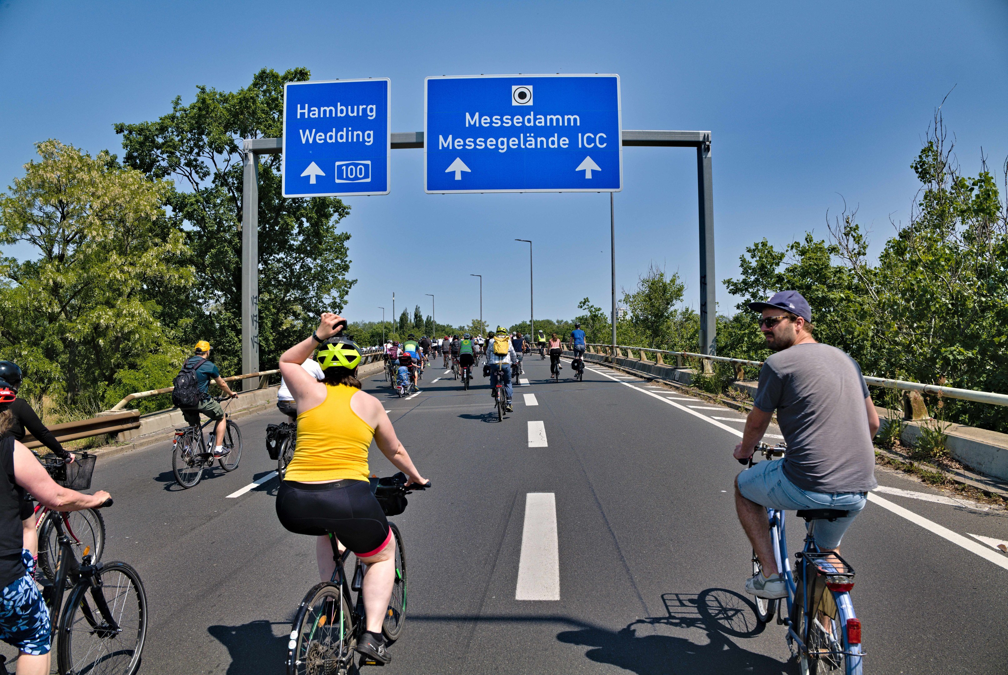 Gruppe von Radfahrern mit Helmen auf einer Straße mit einer Begrenzung auf einer Seite und Bäumen auf der anderen, Laternen im Hintergrund, unter einem klaren blauen Himmel, mit einem Schild, das eine Radtour in Hamburg anzeigt.