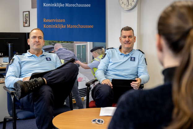 Zwei Polizeibeamte sitzen an einem Tisch mit einer Frau, umgeben von Dokumenten und Gegenständen, mit einer Wanduhr und einem Banner im Hintergrund und einem zweiten Tisch mit einem Monitor und Peripheriegeräten links daneben.