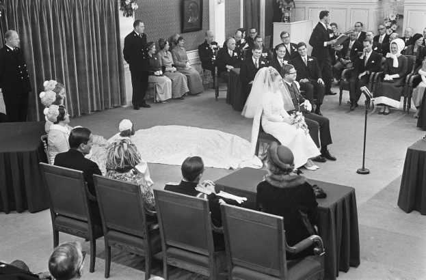 Schwarzes und weißes Foto einer Hochzeitszeremonie mit sitzenden und stehenden Menschen, einem Mann mit Mikrofon und einer Frau mit einem Strauß, mit einem Podium, Blumenschmuck, einem Vorhang, einem gerahmten Foto an der Wand und einer Tür im Hintergrund.