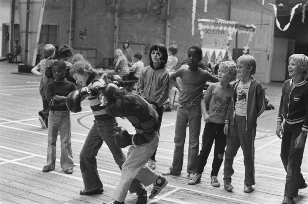 Eine Gruppe von Kindern beim Basketballspielen auf einem Platz, mit zwei streitenden Personen in der Mitte und anderen auf Bänken im Hintergrund, dargestellt in Schwarz-Weiß.