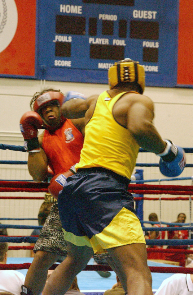 Zwei Boxer in einem Ring, umgeben von einer Menge, mit einer Tafel im Hintergrund, die Text und Logos anzeigt.