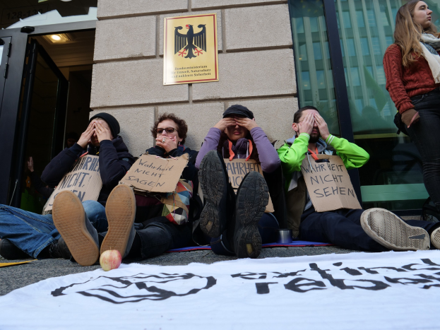 Eine Gruppe von Menschen sitzt vor einem Gebäude auf dem Boden und hält Schilder, mit einem Banner auf der Straße und einer Frau rechts daneben, bei einer Protestaktion gegen eine Regierungsentscheidung.