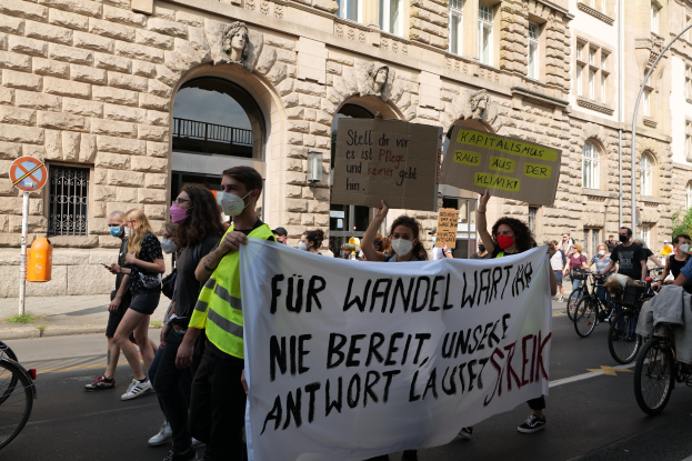 Eine Gruppe von Menschen, die auf einer Straße marschieren, einige halten Schilder und andere fahren Fahrräder, im Hintergrund ein historisches Gebäude mit Rundbogenfenstern, Säulen und Skulpturen.