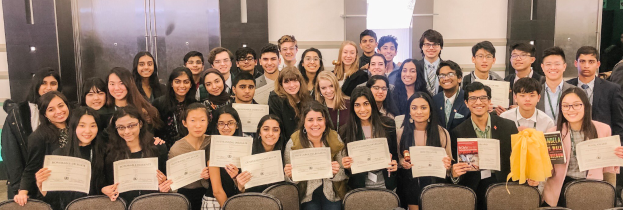 Eine Gruppe von Menschen hält Urkunden in den Händen und posiert für ein Foto bei einer Abschlussfeier, mit Stühlen und einer Wand im Hintergrund.
