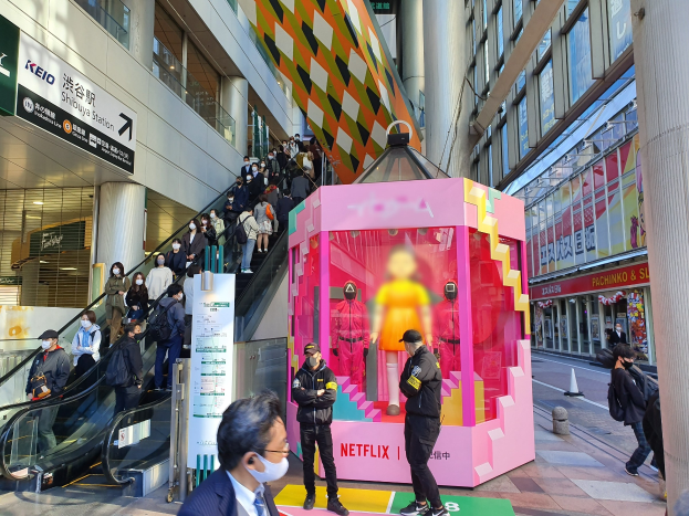 Gruppe von Menschen vor einem Glaswandgebäude mit Texttafeln, einige tragen Masken, in der Nähe einer Rolltreppe mit Menschen und einer pinken Box in der Mitte mit Schaufensterpuppen und dem Text "Netflix's new Netflix Booth in Tokyo".