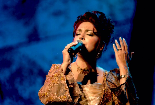 Frau in goldenem Kleid, das in ein Mikrofon singt, auf einer Bühne beim Eurovision Song Contest mit blau-schwarzem Hintergrund.